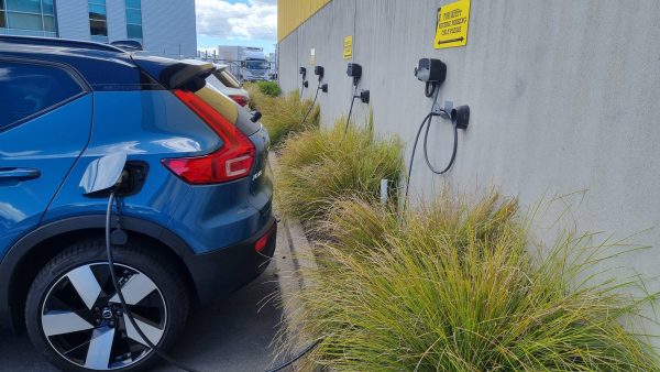 Blue electric vehicle plugged into a wall-mounted EV charging station in a parking lot, with other charging points and signage visible along the concrete wall