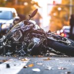 A heavily damaged motorcycle lying on its side in the middle of a city road after a collision. The scene includes cars stopped in the background, scattered debris, and blurred figures of pedestrians nearby, with autumn leaves scattered on the ground and a warm, golden light in the distance.