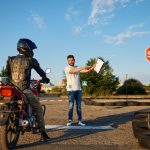 A motorcycle instructor holding a clipboard provides guidance to a learner wearing protective riding gear and sitting on a red motorcycle. The training area features a stop sign, tire barriers, and open space under a clear blue sky, creating a safe environment for learning.