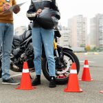 A motorcycle training instructor holding a clipboard gives guidance to a learner motorcyclist. The learner, dressed in a leather jacket and jeans, holds a helmet while standing beside a black motorcycle. Red safety cones surround them on the pavement, with apartment buildings visible in the background.