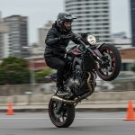 A motorcyclist wearing a black helmet and black riding gear performs a wheelie on a powerful sportbike in an urban setting. The front wheel is lifted off the ground as the rider skillfully balances the bike, with a backdrop of tall buildings and blurred city scenery.