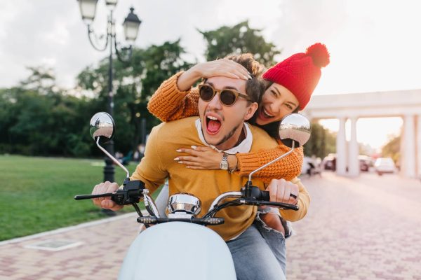 A cheerful couple riding a white scooter, with the man in sunglasses laughing and the woman in a red beanie and orange sweater playfully hugging him from behind, enjoying a fun moment outdoors on a bright day.
