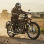 A rider wearing a helmet and leather jacket rides a vintage motorcycle on a dusty dirt road during golden hour. The countryside scenery features open fields and scattered trees in the distance, with warm sunlight creating a nostalgic ambiance. The motorcycle’s classic design is highlighted, capturing a timeless moment of adventure and freedom.