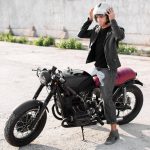 A young man wearing a leather jacket and jeans adjusts his helmet while standing next to a matte black vintage-style motorcycle with a red seat. The setting is an urban outdoor area with a concrete surface and a white wall in the background, exuding a retro and modern biker aesthetic.
